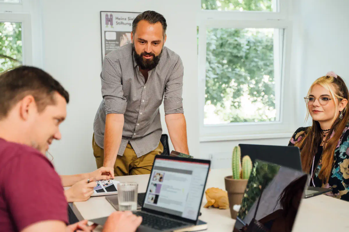 Drei Mitglieder eines Marketingteams besprechen eine Strategie an einem Tisch mit Laptops